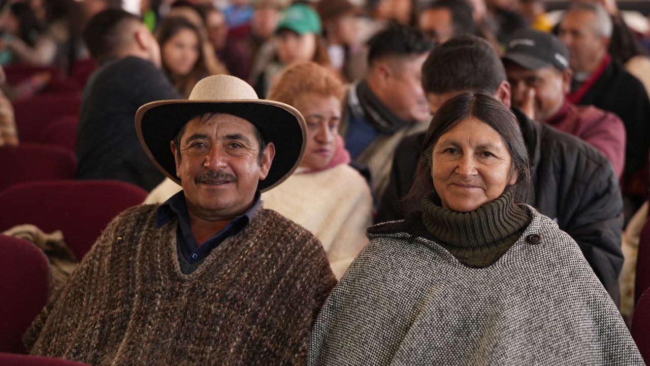En la imagen se ve a dos personas sentadas una al lado de la otra en lo que parece ser un auditorio. La persona de la izquierda lleva un sombrero de ala ancha y un jersey, mientras que la de la derecha lleva un chal y un jersey de cuello alto. Ambos sonríen y parecen divertirse. El fondo está lleno de gente, lo que sugiere que están asistiendo a un evento o reunión. El ambiente parece informal y relajado.